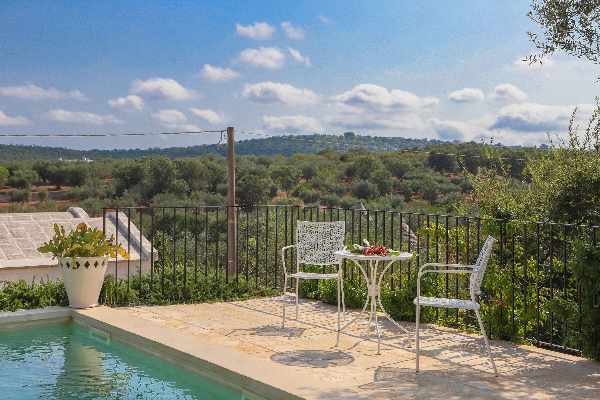 A poolside area with a table and chairs, surrounded by a fence, overlooking a landscape with trees and hills.