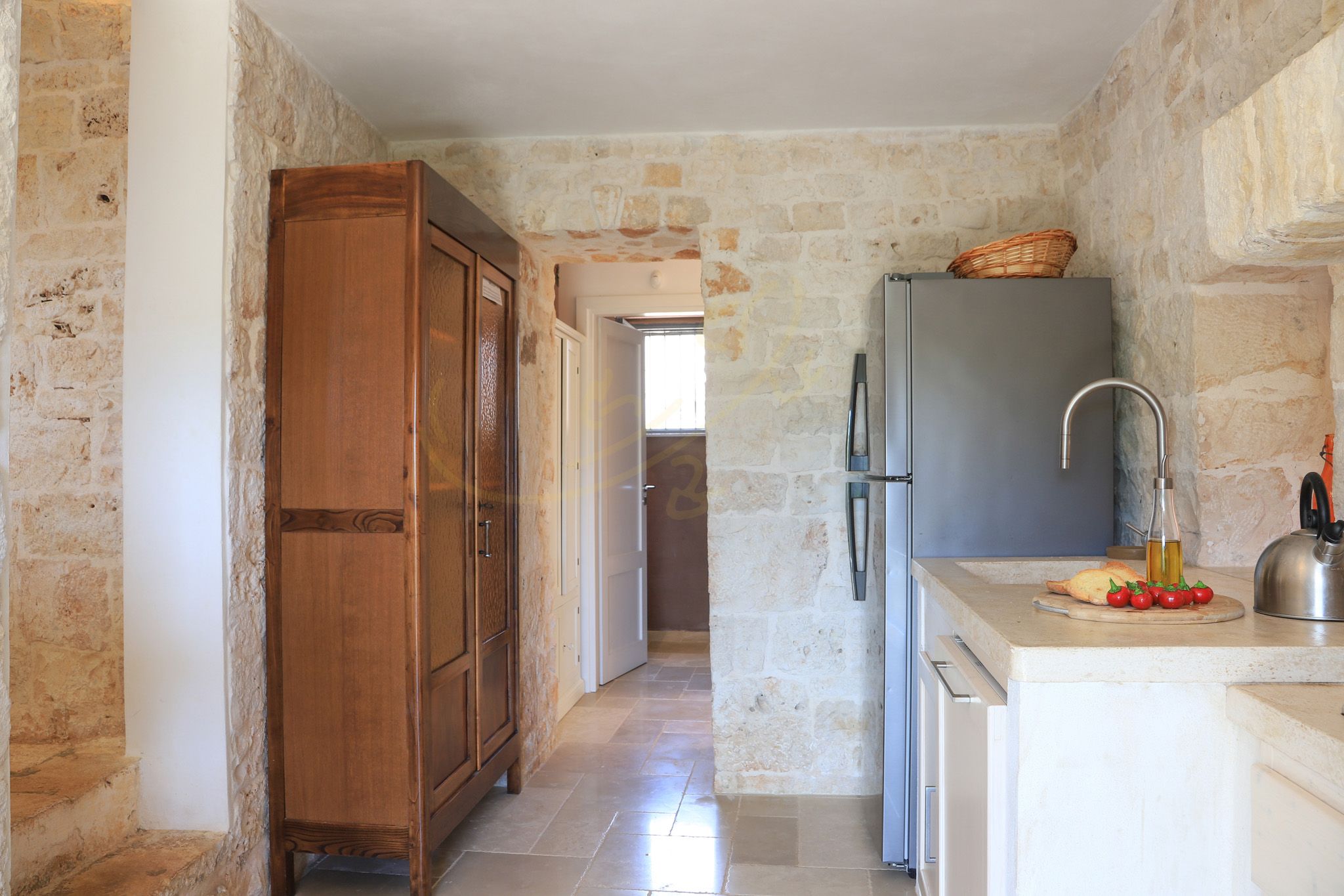 A narrow kitchen with stone walls, a wooden cabinet, a refrigerator, and a countertop with a bottle and tomatoes.