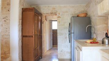 A narrow kitchen with stone walls, a wooden cabinet, a refrigerator, and a countertop with a bottle and tomatoes.