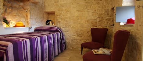 Une pièce aux murs de pierre, un lit avec une literie à rayures, une chaise rouge et une petite étagère sur laquelle se trouve un chapeau rouge.