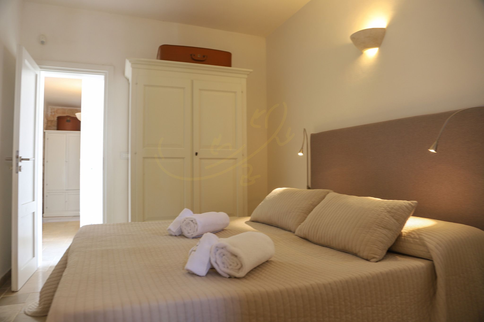 A bedroom with a bed, two rolled towels, a wardrobe, and a wall-mounted light fixture.