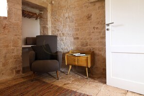 A room with a stone wall, a grey armchair with a cushion, a wooden side table with books, and a white door. - TD Casale Terranova Stone Farmhouse with Pool (Monopoli)