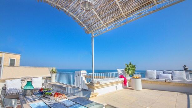 Terraço com mesa posta para refeições, área de estar com almofadas e vista para o mar.