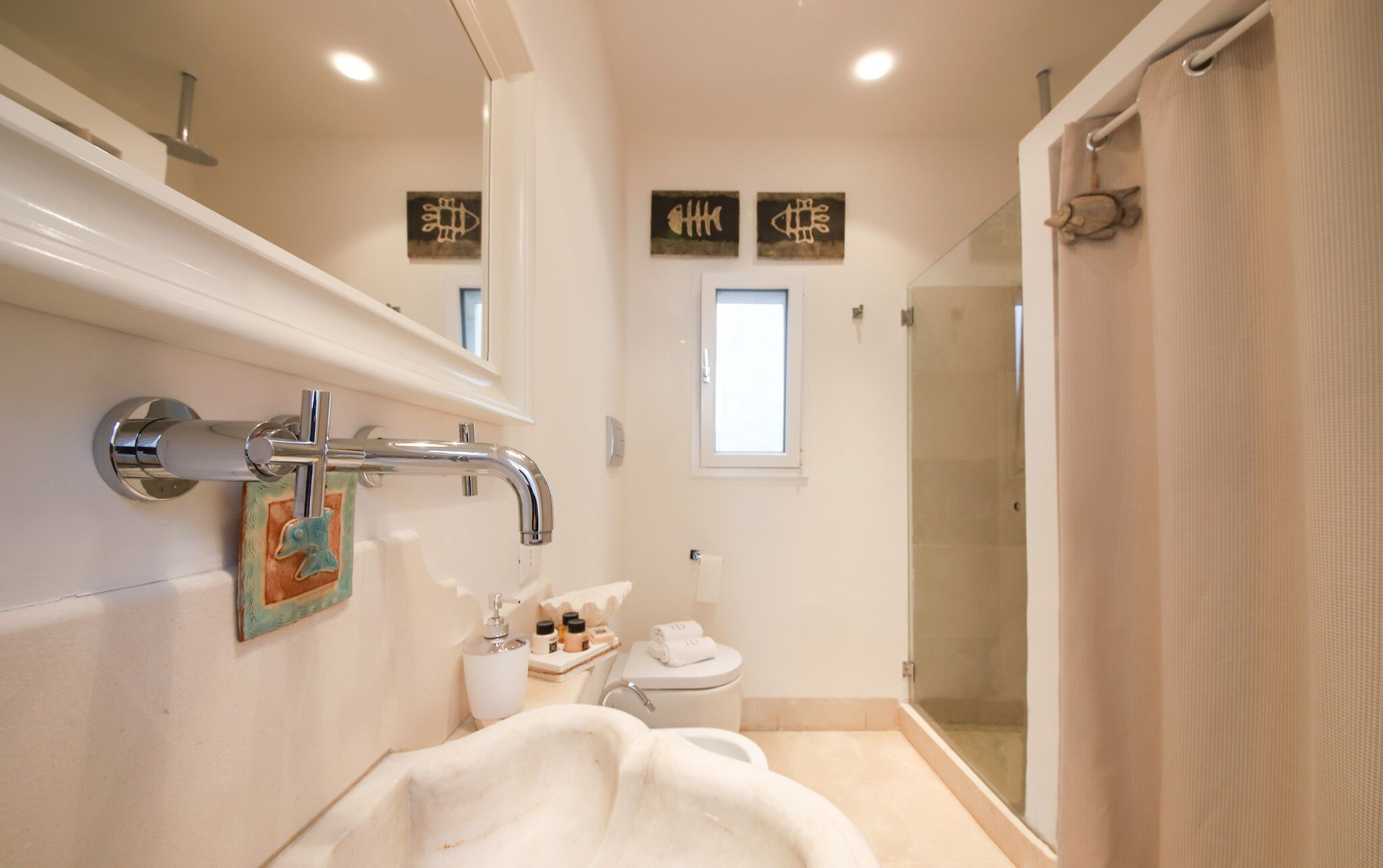 A modern bathroom with a bathtub, toilet, and shower. There are wall-mounted artworks and a window above the toilet.