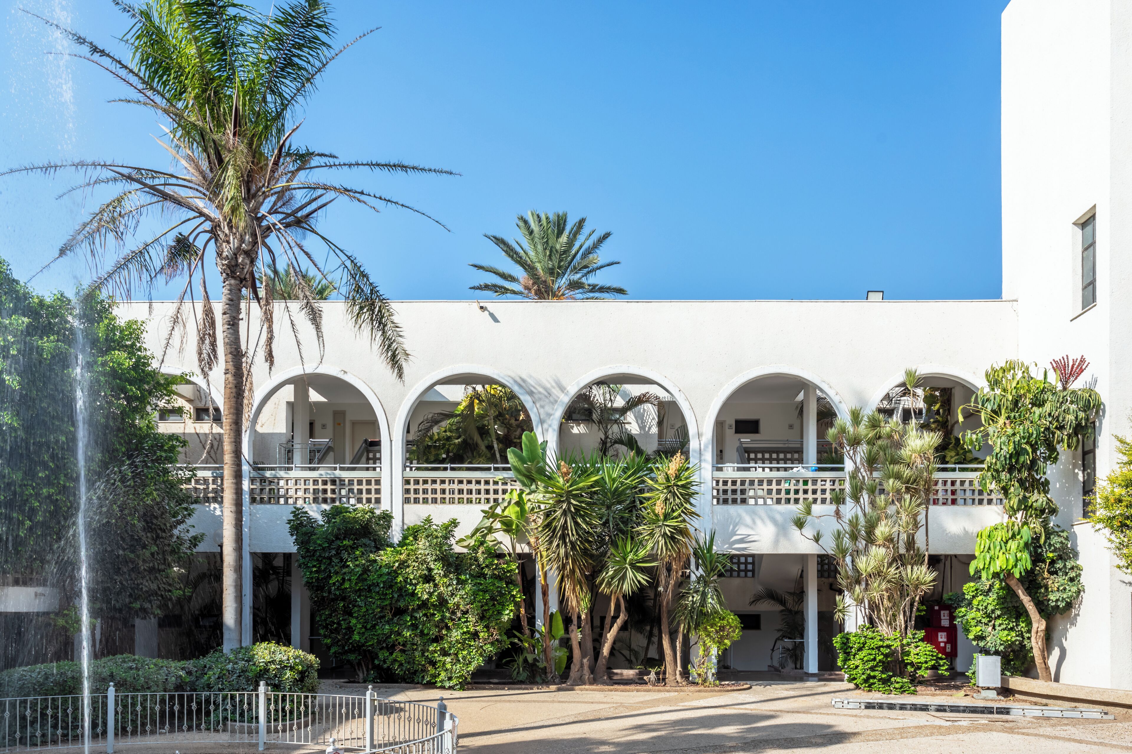 A building with multiple arches, a palm tree, and a fountain.