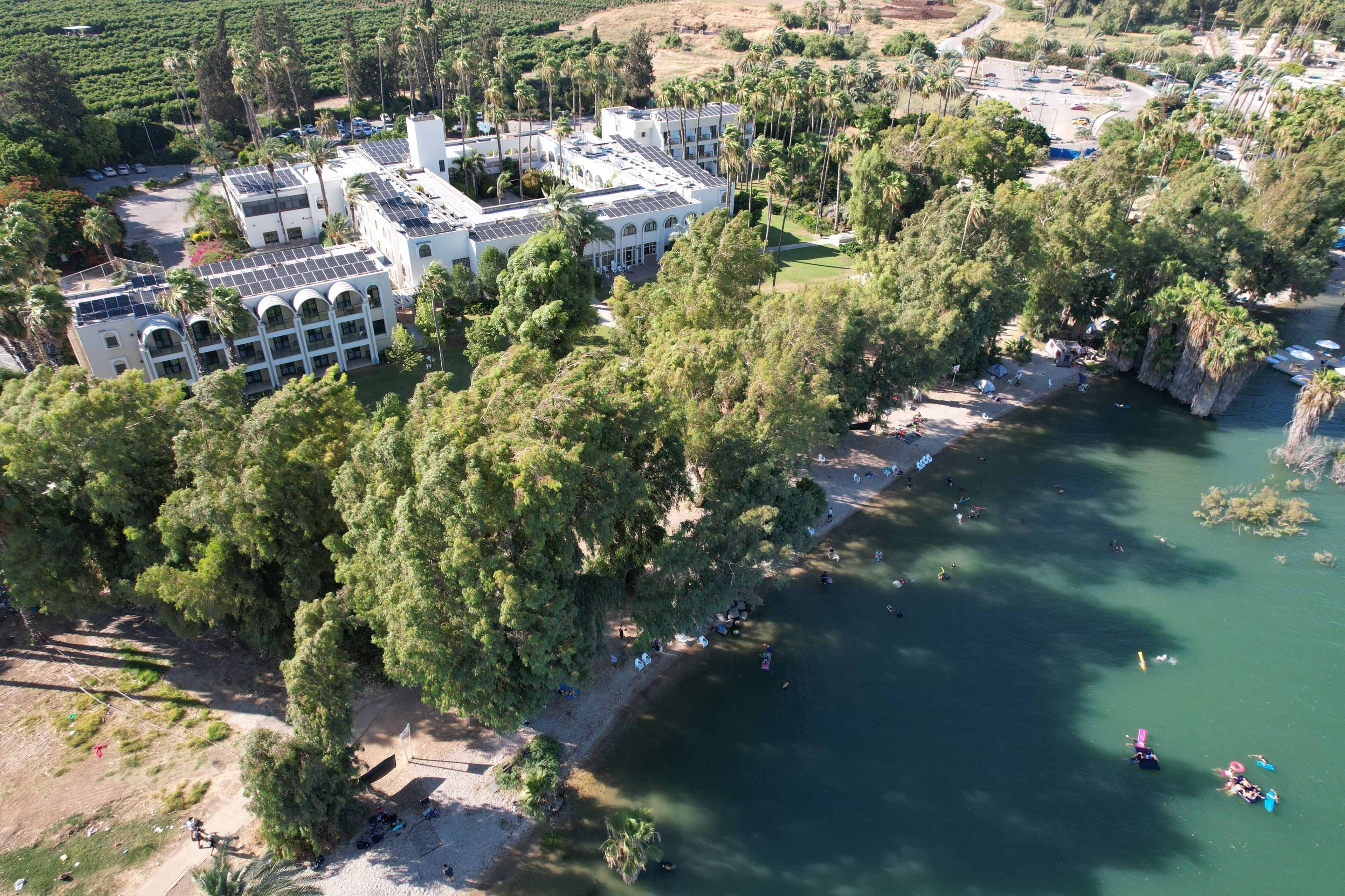 Vista aérea de un hotel con varias construcciones, rodeado de abundante vegetación y una zona de playa con gente y barcos.