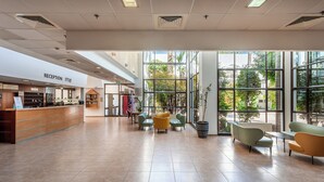 A reception area with a reception desk, seating area with chairs, large windows, and indoor plants. - HI Karei Deshe Hostel (Emek HaYarden)