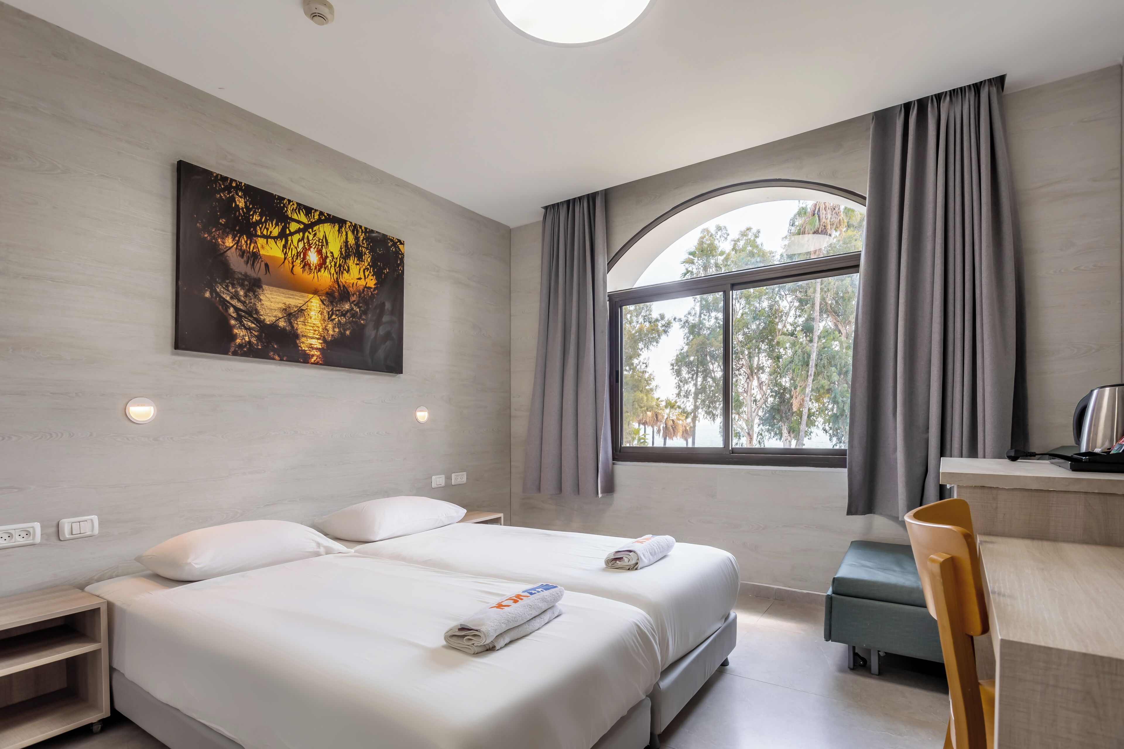 A hotel room with two beds, a desk, a chair, a large window with curtains, and a painting on the wall.