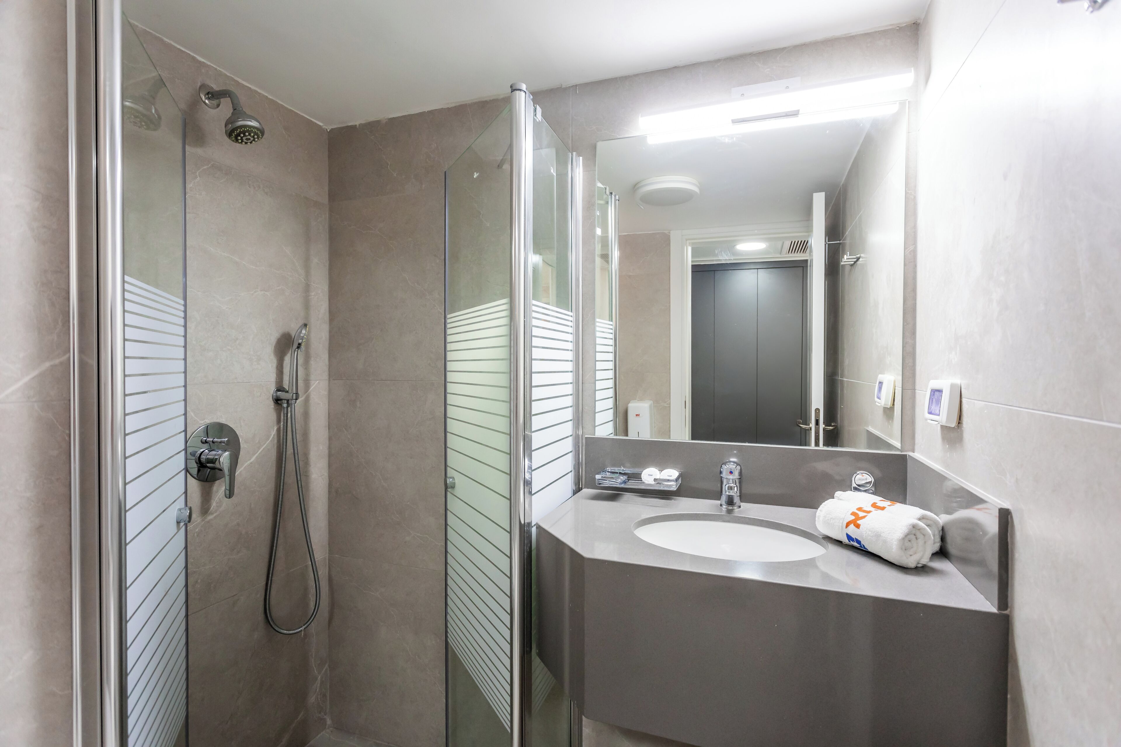Un baño moderno con ducha acristalada, lavamanos con toalla y espejo que refleja el cuarto.