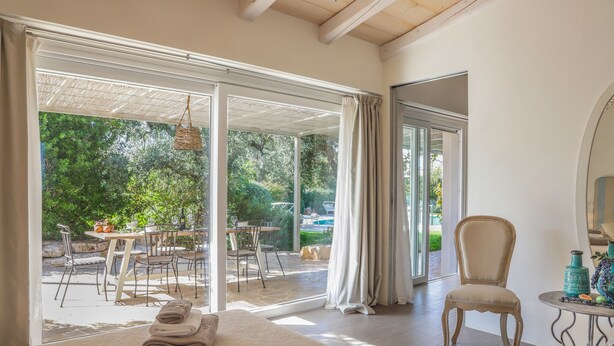 Ein Zimmer mit einem Bett, einem Sessel, einem Tisch und Blick auf eine Terrasse mit Tisch und Stühlen.
