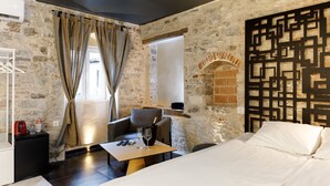 Una habitación con pared de piedra, una cama, una mesa con vasos y una botella, y una silla.