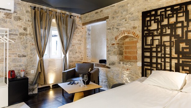 A room with a stone wall, a bed, a table with glasses and a bottle, and a chair. - Palace Luxury Room (Split)