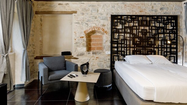 Una habitación de hotel moderna con pared de piedra, cama, sofá gris, pequeña mesa con botella y vasos, y cabecero decorativo negro.