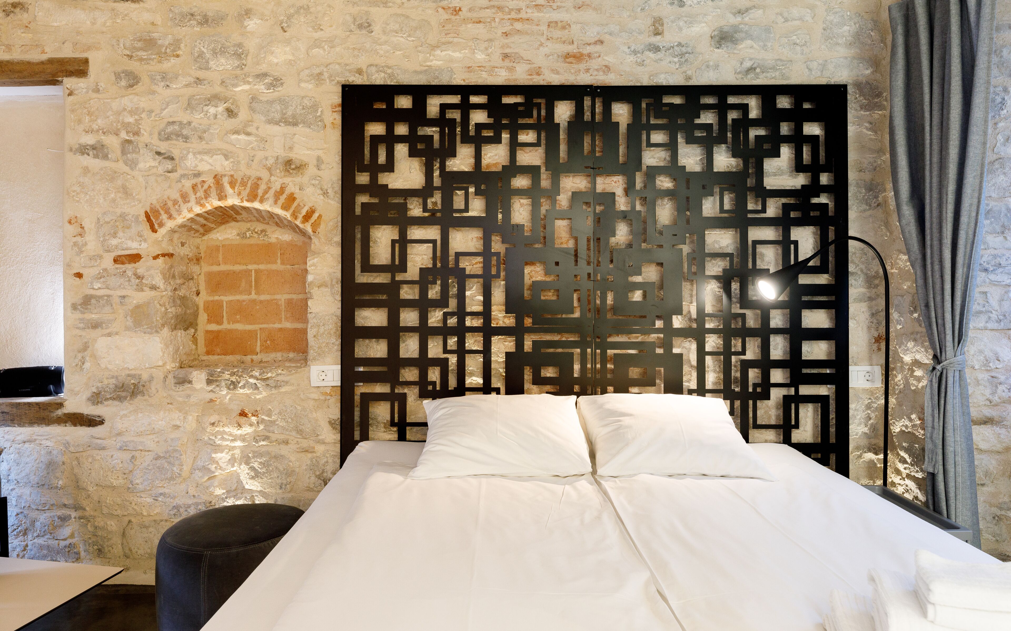 A bedroom with a stone wall, a decorative screen, a bed with white linens, and a grey curtain.