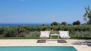 Zona de piscina con dos sillas reclinables blancas y una mesita, con vista a un cielo azul despejado y al océano.