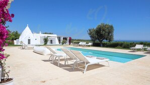 Un área de piscina con sillones de descanso, un edificio blanco con techo cónico y vistas al mar.