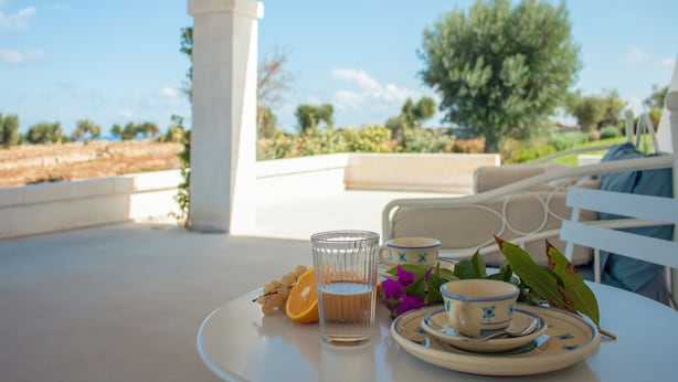 Una mesa con un vaso de agua, una taza y un plato con comida y flores en una terraza con vistas a árboles y un cielo despejado.