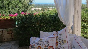 Un balcon avec un rideau blanc, une vue sur la verdure et des bâtiments éloignés, et un lit agrémenté d’oreillers à motifs.