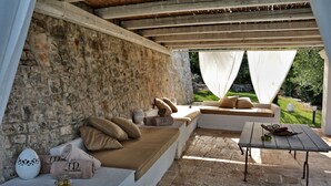 Un espace extérieur aménagé avec des murs en pierre, des bancs rembourrés et une table sur laquelle se trouve un panier de fruits.