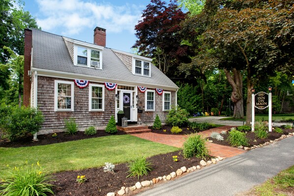 A two-story house with a brick chimney, white trim, and a front porch. There is a driveway leading to the house, and a sign indicating the address. - Inn on Main (Yarmouth Port)