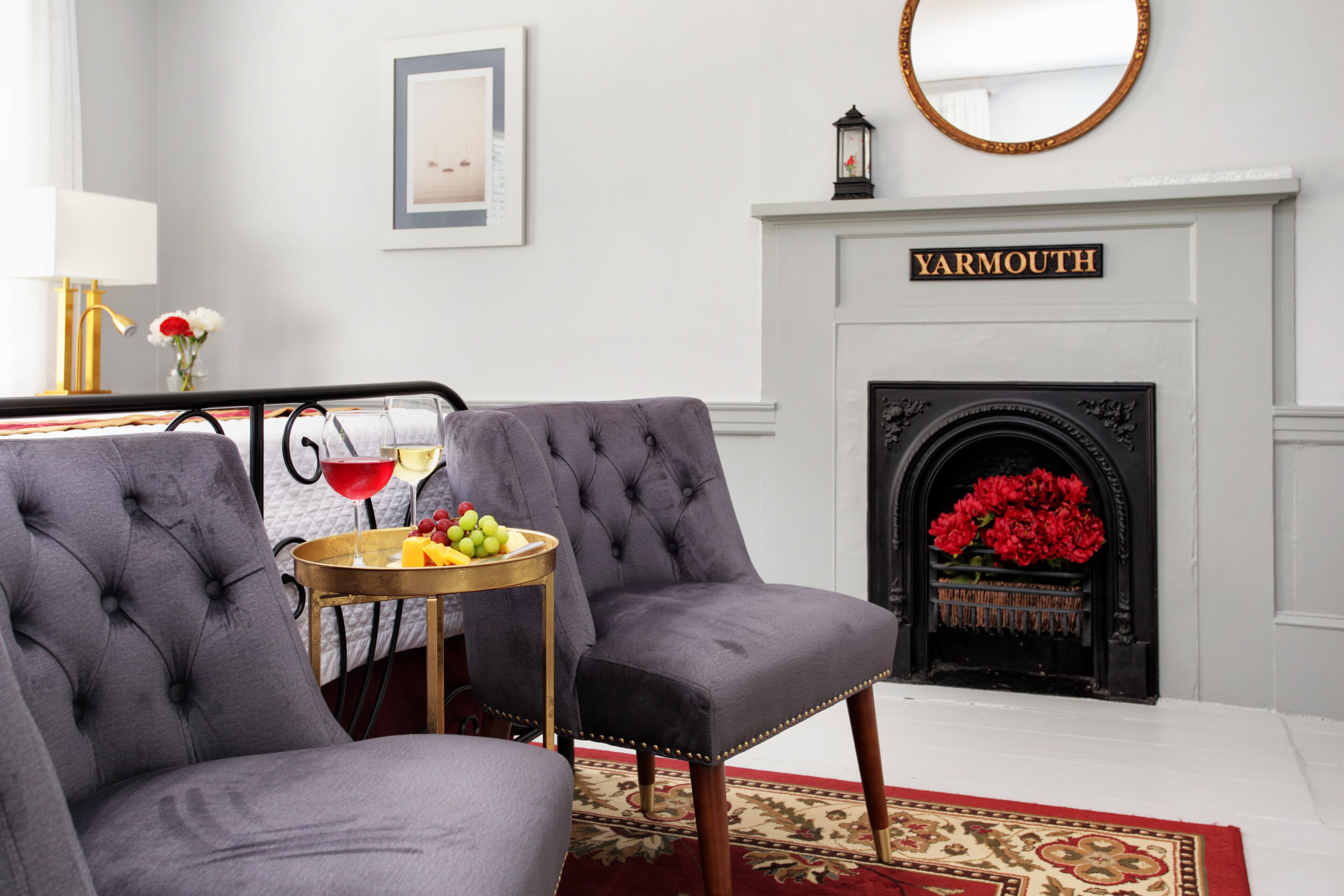 A living room with two grey upholstered chairs, a small wooden side table with fruit and a drink, a fireplace with a 'YARMOUTH' sign, and a red and gold patterned rug.