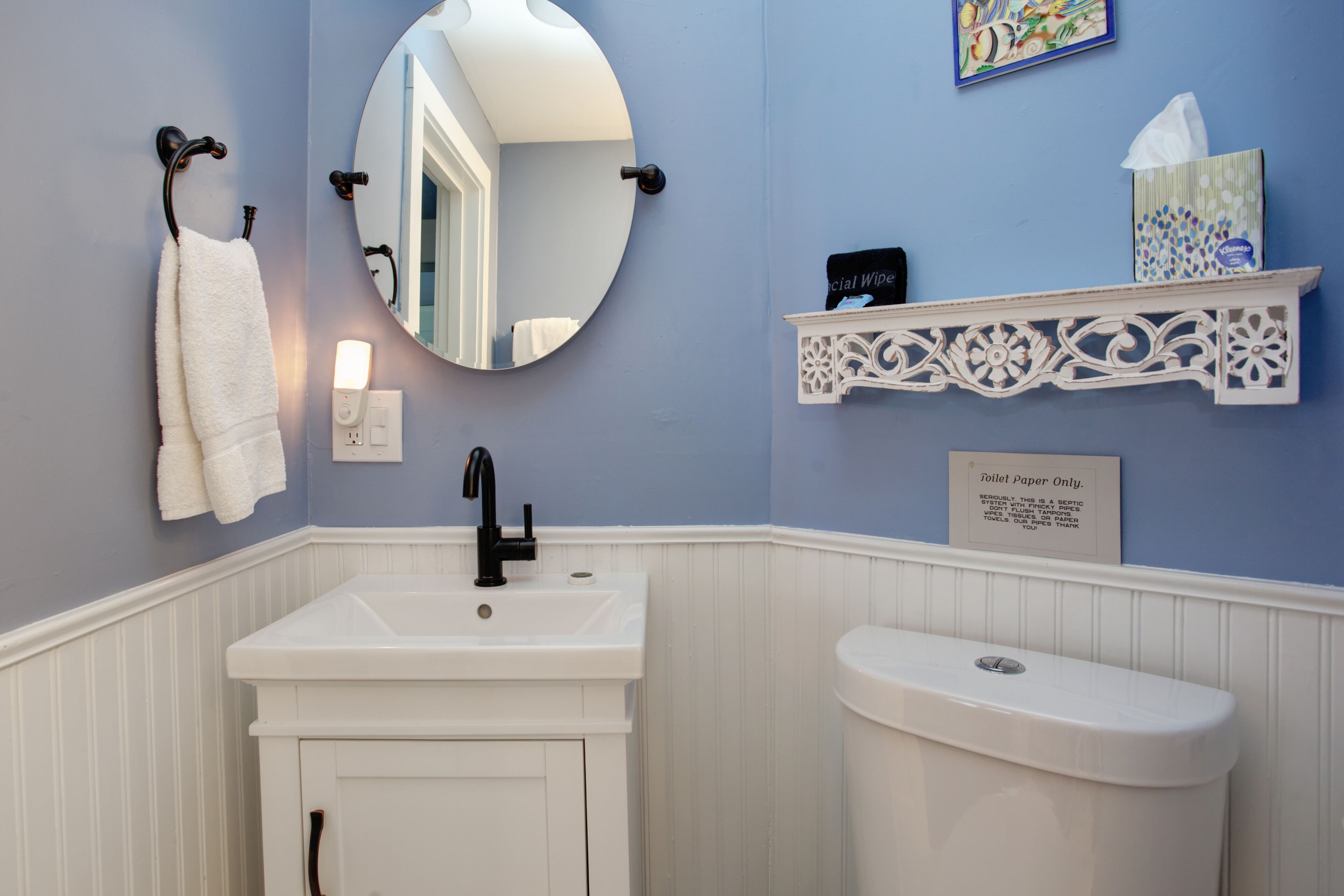 A bathroom with a white sink, black faucet, and a white toilet. There is a mirror above the sink, a towel rack with a white towel, and a decorative shelf with a tissue box and a sign that says "Toilet Paper Only."