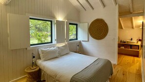 A bedroom with a wooden ceiling, a bed with white bedding, a red and black rug, and a window with a view of greenery.
