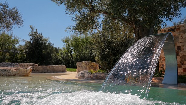 A water feature with a curved waterfall in a landscaped area with rocks and trees. - TD Trulli di Montaltino with Stunning Huge Pool (Polignano a Mare)