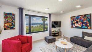 Un salon moderne avec un canapé rouge, un canapé gris et une table basse blanche. Deux tableaux abstraits ornent les murs, une grande fenêtre offre une vue dégagée et un téléviseur à écran plat est fixé au mur.