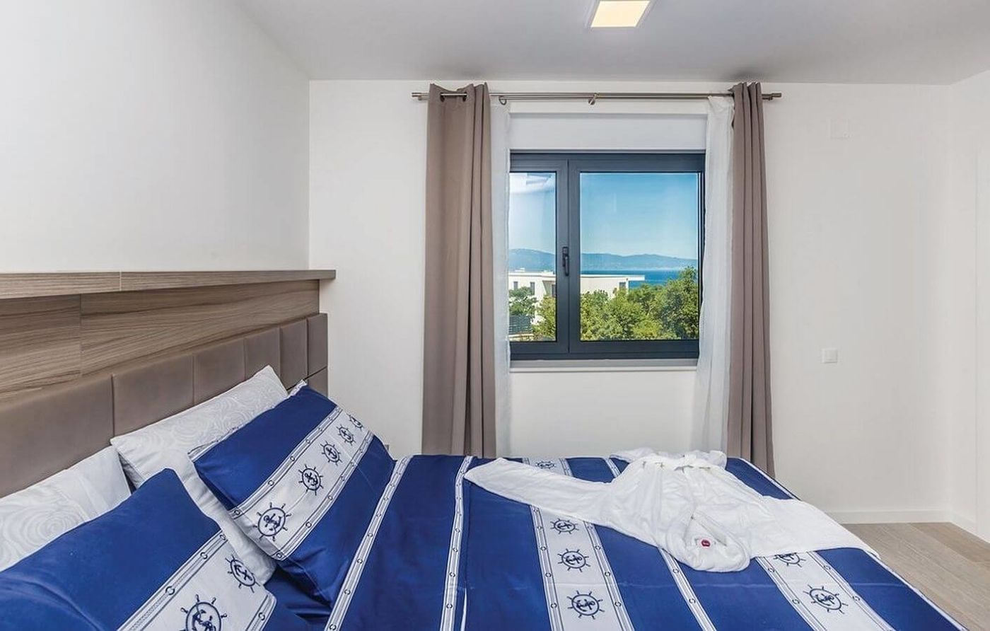 Ein Schlafzimmer mit Bett, einem Fenster mit Vorhängen und Blick auf Gebäude und Grünflächen.