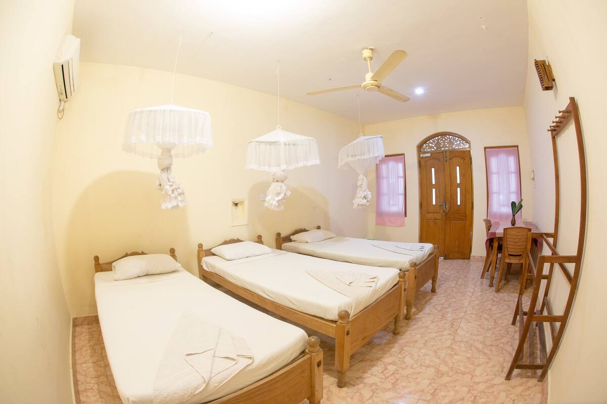 Una habitación con tres camas de madera, un ventilador de techo, una mesa de madera con sillas y una puerta con arco decorativo.