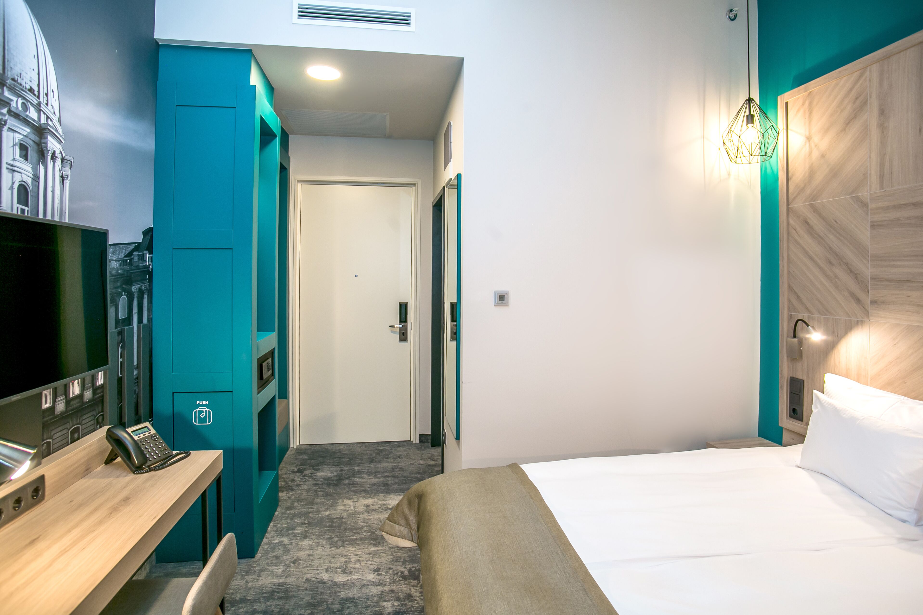 A hotel room with a bed, a desk with a telephone, a wall-mounted TV, and a door leading to a hallway.