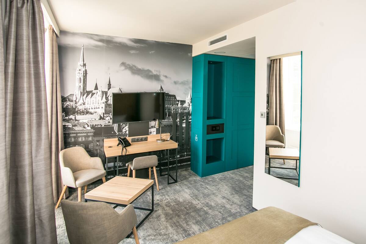Hotellihuone, jossa on suuri sänky, työpöytä tietokoneella, kaksi tuolia, televisio ja seinämaalaus, jossa on esitetty kaupunkimaisema.