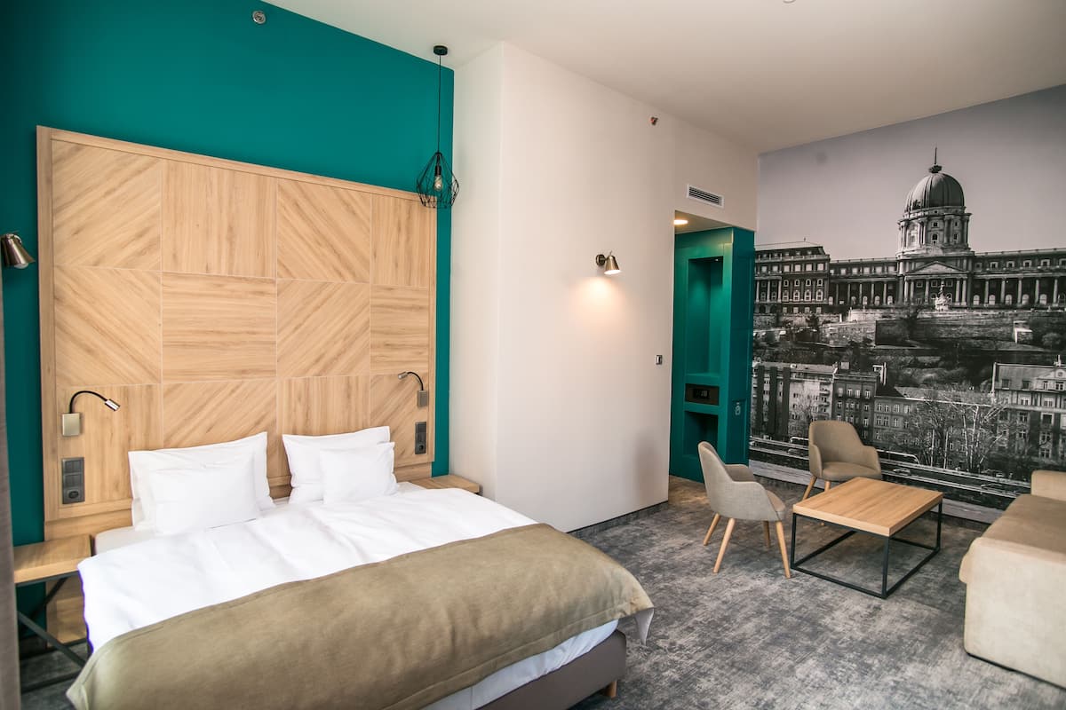 Hotellihuone, jossa on suuri sänky, puinen pääty, oleskelualue pöydällä ja tuoleilla sekä seinällä kaupunkimaisemaa esittävä seinämaalaus.