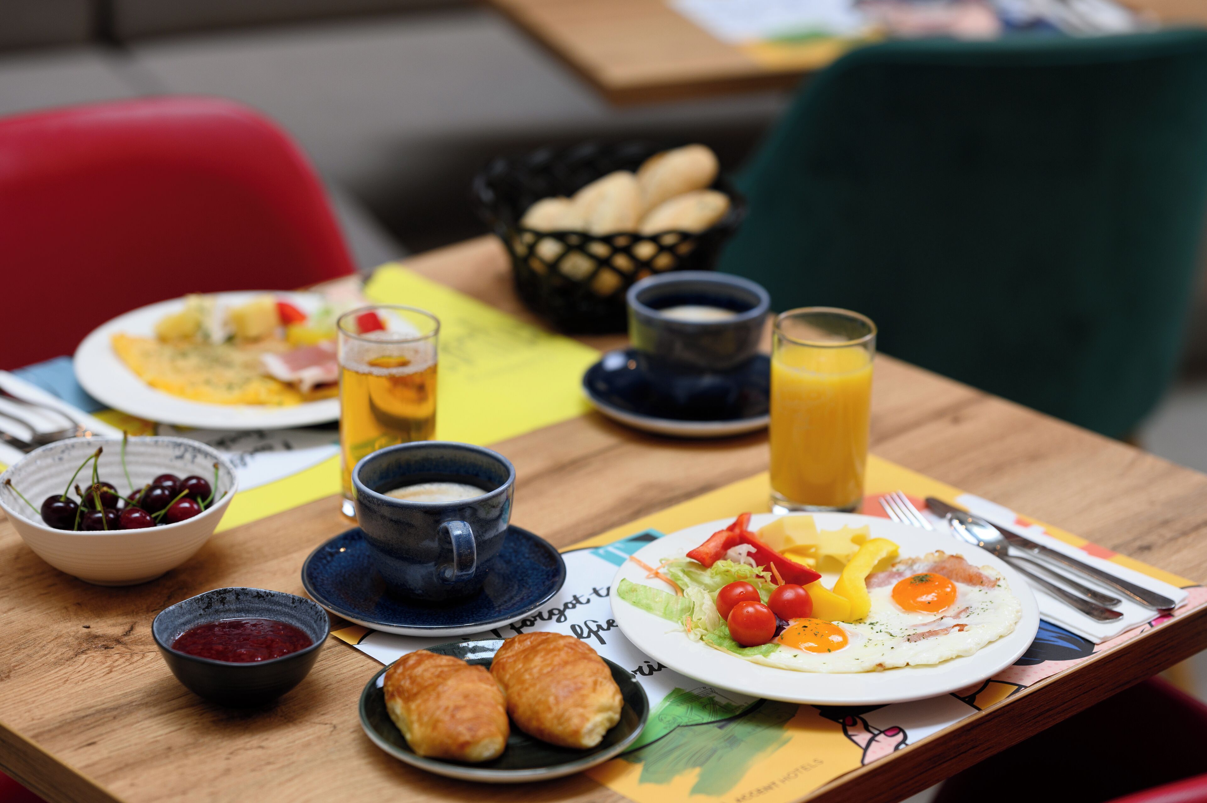 Une table de petit-déjeuner avec des assiettes remplies de nourriture, un bol de cerises, une tasse de café et un verre de jus d’orange.