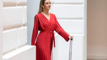 A person in a red dress standing next to a suitcase in a hallway with white walls and a tiled floor.