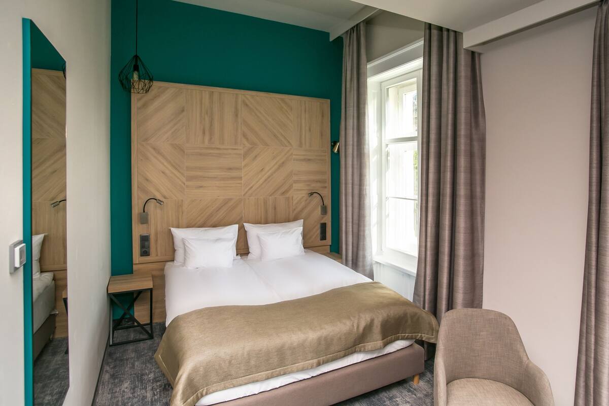 Hotellihuone, jossa on suuri sänky, puinen pääty, yöpöytä, tuoli ja ikkuna, jossa on verhot.