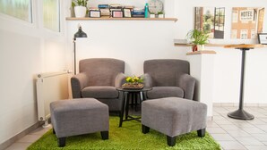 Un salon moderne avec deux fauteuils gris, une petite table ronde ornée d’un vase de fleurs, un radiateur, une lampe sur pied et un tapis vert.