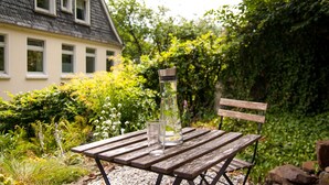 Une table en bois, sur laquelle sont posés un verre et une carafe, est placée à l’extérieur, avec une maison et de la verdure en arrière-plan.