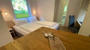 Une chambre d’hôtel avec une table en bois, un lit avec du linge de maison blanc, une chaise et une fenêtre donnant sur un paysage verdoyant.