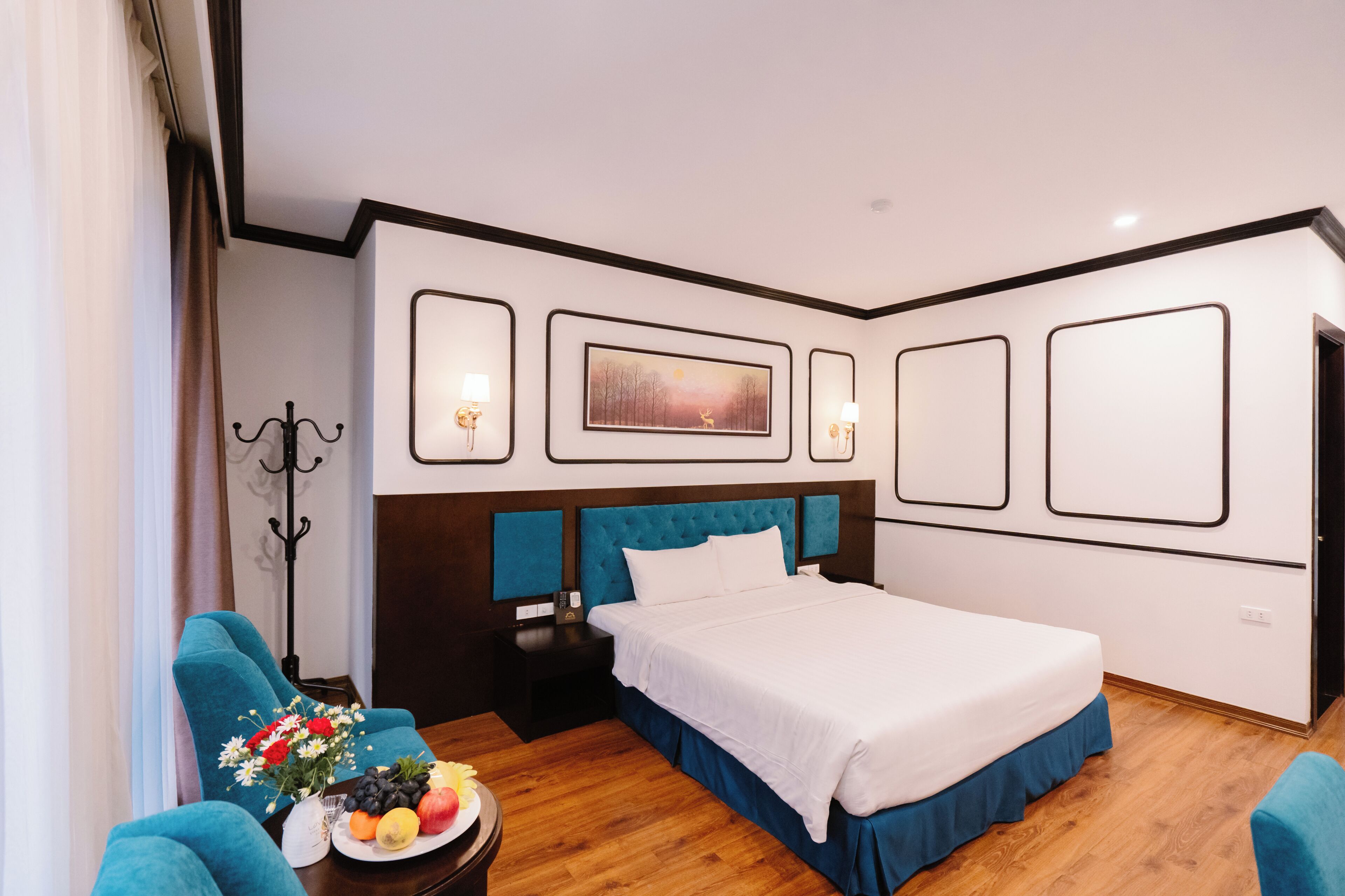 Une chambre d’hôtel moderne avec un grand lit, une table de chevet, un fauteuil bleu, une table basse avec des fruits et un tableau au mur.