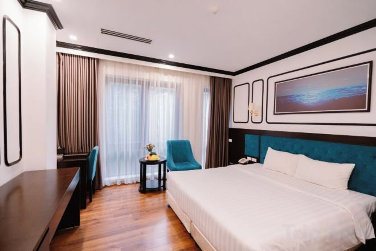 A hotel room with a bed, a desk, a chair, a small table with a lamp, and a window with curtains.