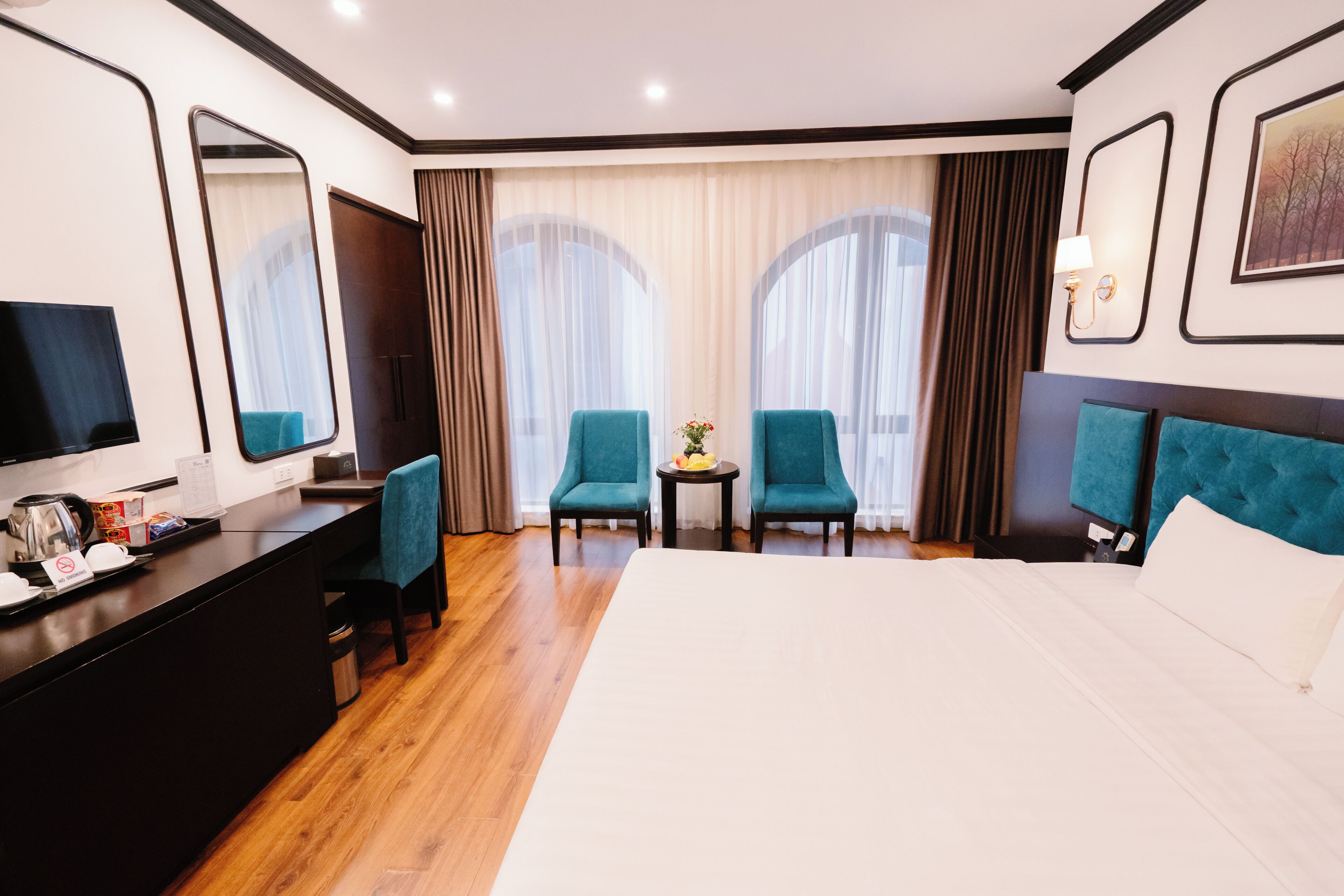 Een hotelkamer met een bed, twee stoelen, een bureau met een televisie en een kleine tafel met een bloevas.
