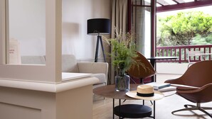Un salon moderne avec un canapé, une table basse ronde ornée d’un vase et d’un chapeau, un lampadaire et une vue sur la verdure à l’extérieur.