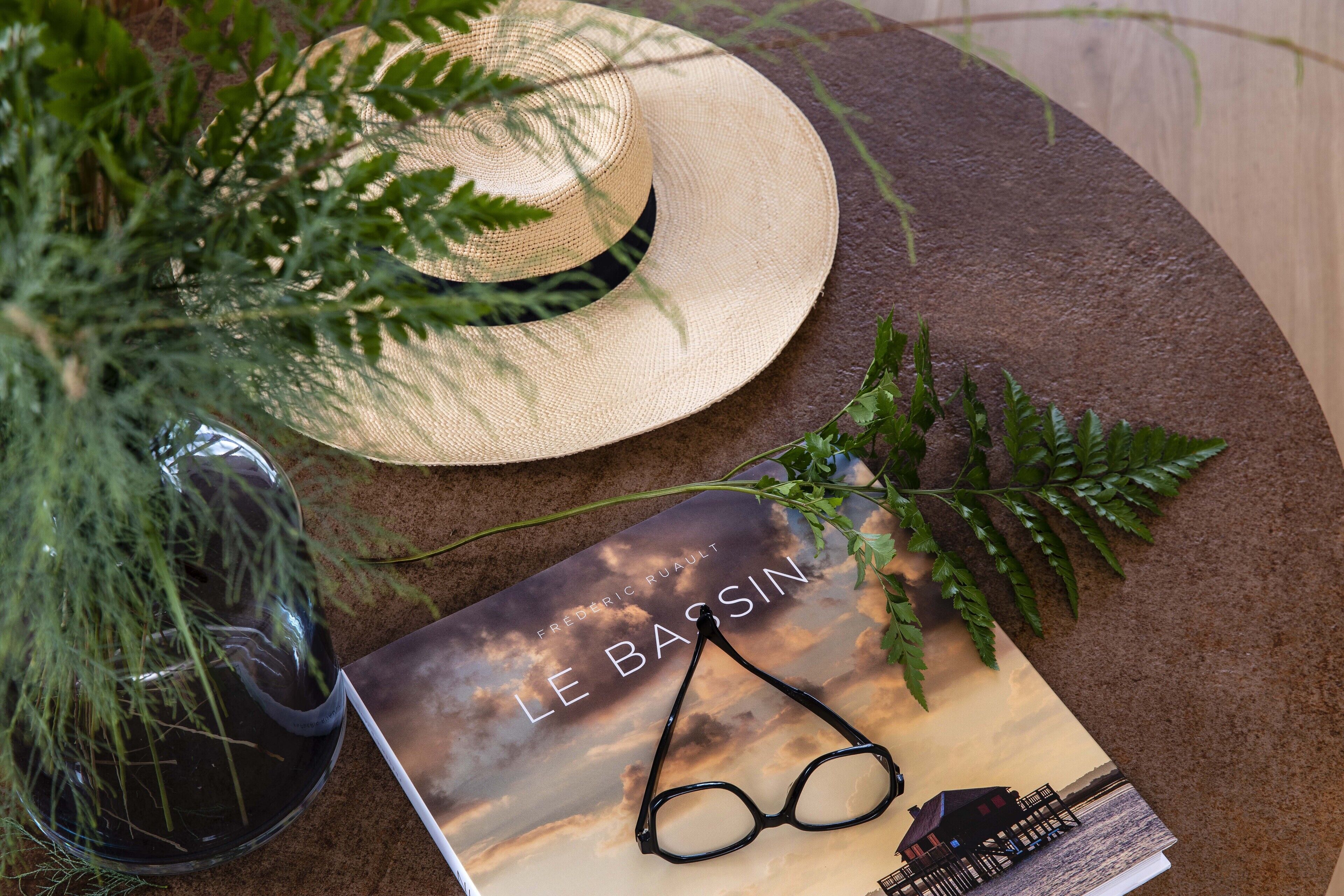 A book titled "Le Bassin" with a pair of glasses on top, placed on a textured surface with a straw hat and green foliage nearby.