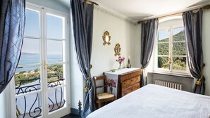 A bedroom with a bed, a dresser, a chair, and a view of the sea through a large window. - Villa Cà Mea (La Spezia)