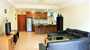 A modern living room with a black leather sofa, a glass coffee table, and a TV on a wooden stand. The kitchen area is visible with wooden cabinets and a refrigerator. - View Talay 2 - 1 bed Jomtien (Pattaya)