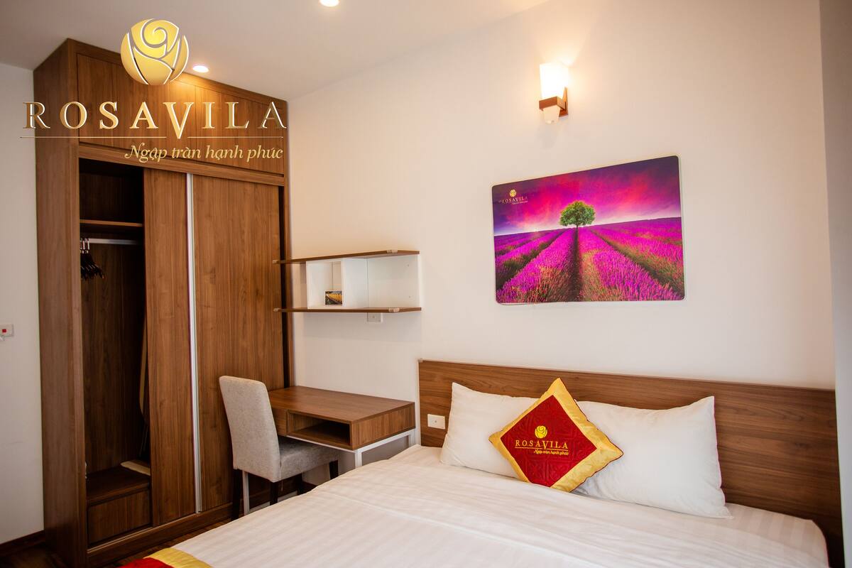 A hotel room with a wooden wardrobe, a desk with a chair, a bed with a pillow, and a wall painting of a field.