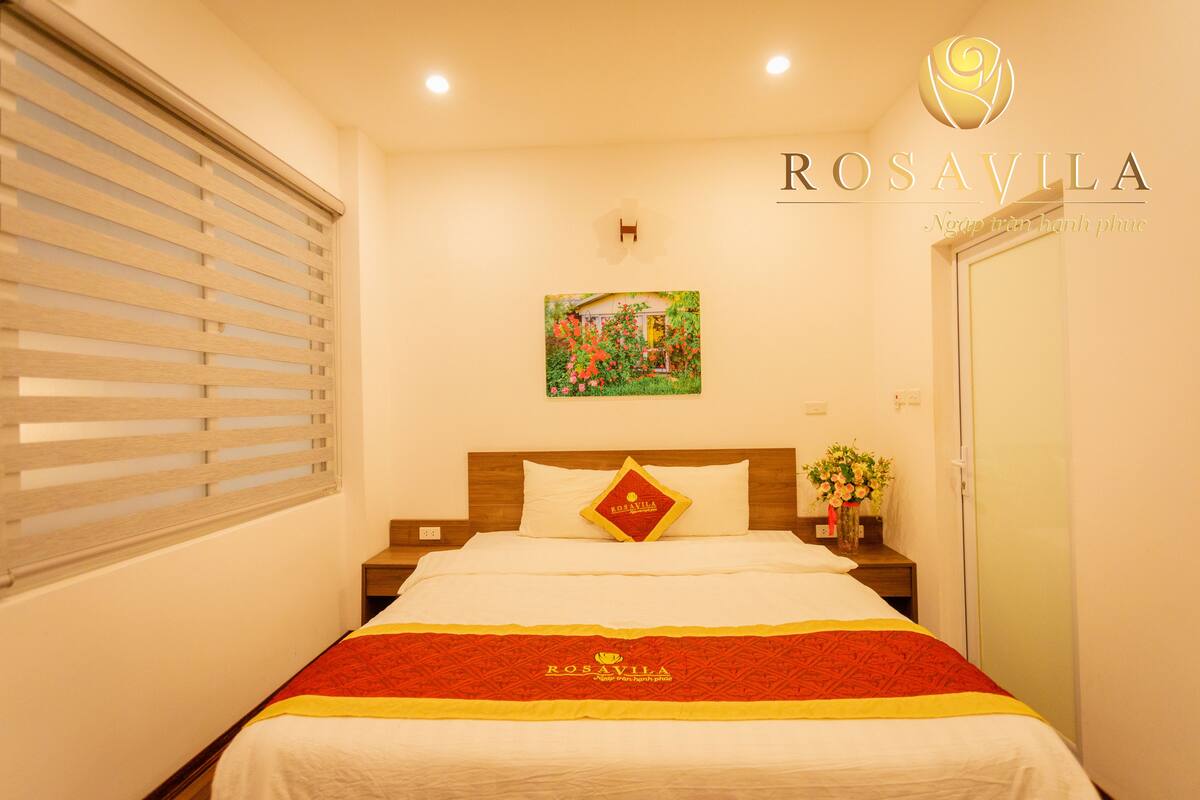A hotel room with a bed, a nightstand, a window with blinds, a painting on the wall, and a door.