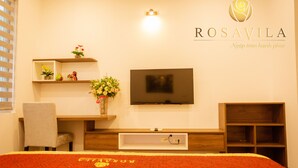 A hotel room with a bed, a TV, a desk with a chair, a shelf with flowers, and a wooden cabinet. - Rosavila (Phan Dinh Phung)